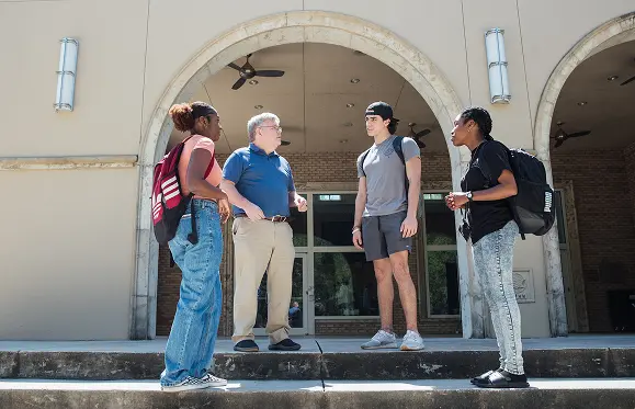 A man speaks with three students carrying backpacks outside a building with arched doorways.
